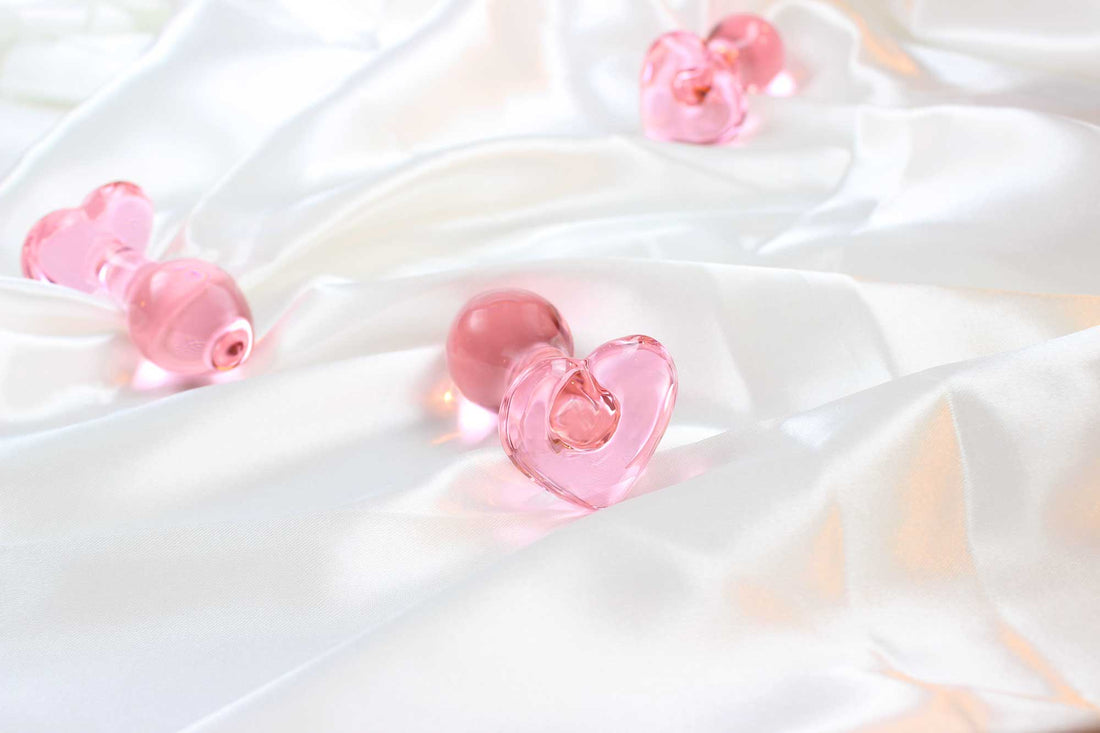 Three pink, glass butt plugs with heart shaped bases on white satin