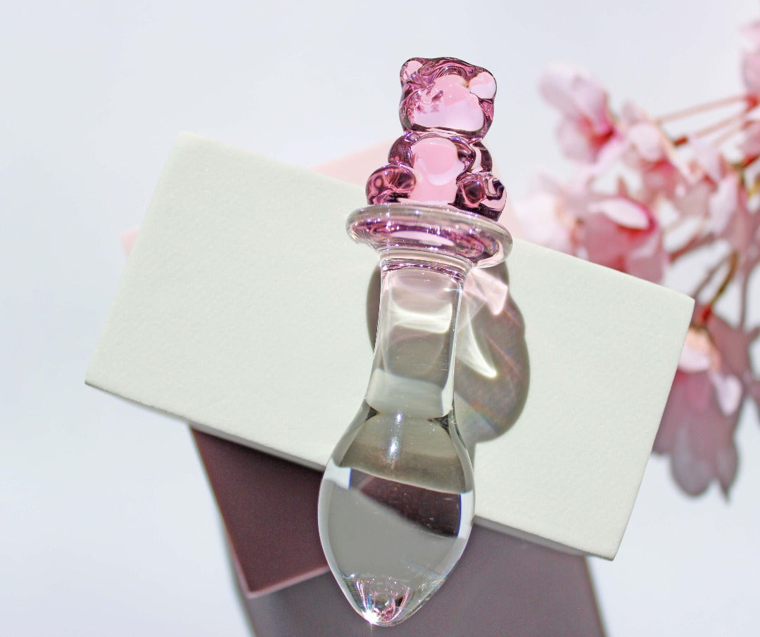 Bear glass butt plug with a bulbed shaft and a pink bear base being leaned against a white cube on a white background with pink flowers.
