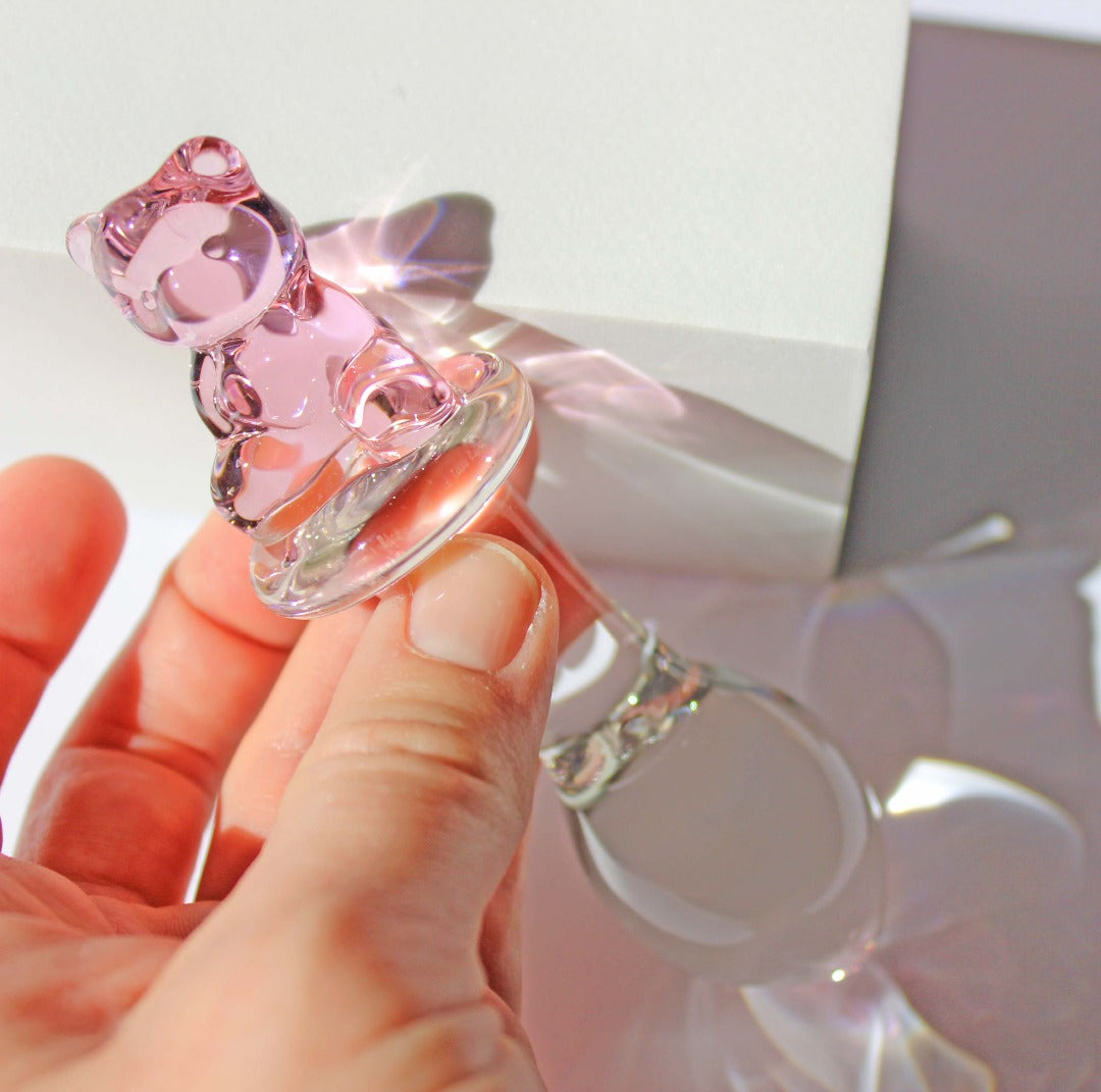 Bear glass butt plug with a bulbed shaft and a pink bear base being held in front of a white cube on a white background.