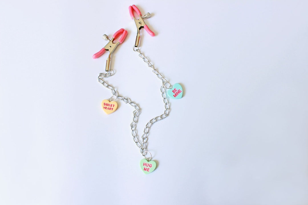 Pink PVC Capped silver tension screw nipple clamps connected by a chain with three candy heart charms on them placed on a white background.