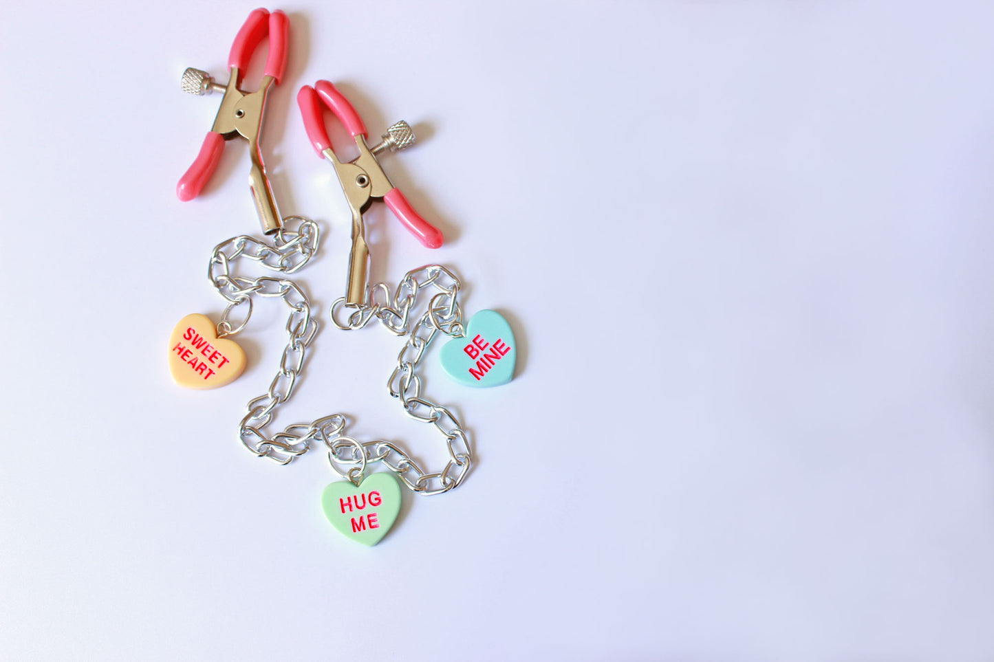 Pink PVC Capped silver tension screw nipple clamps connected by a chain with three candy heart charms on them placed on a white background.