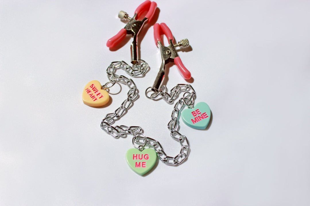 Pink PVC Capped silver tension screw nipple clamps connected by a chain with three candy heart charms on them placed on a white background.