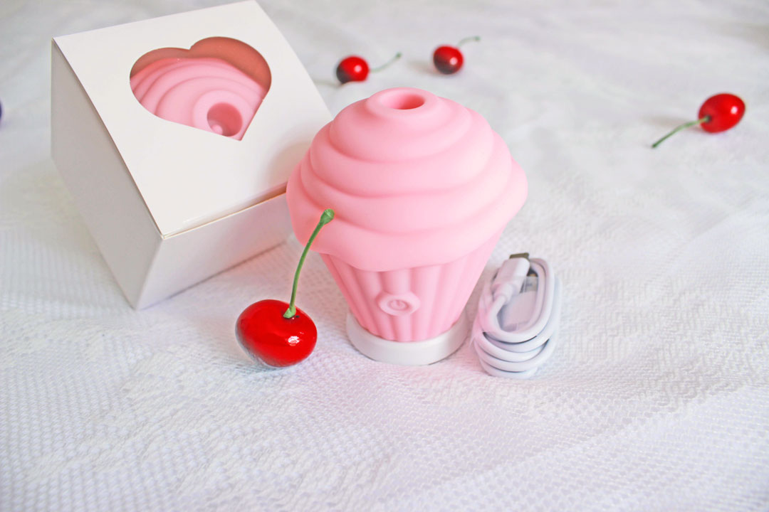 Pink sucking cupcake vibrator next to its usb charger in front of its packaging boxPink sucking cupcake vibrator next to its usb charger in front of its packaging box