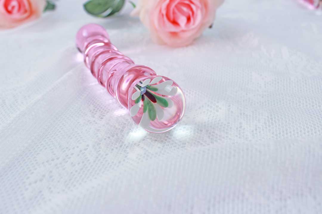 Pink glass ridged dildo with a flower base laid on white lace with pink roses in the background.