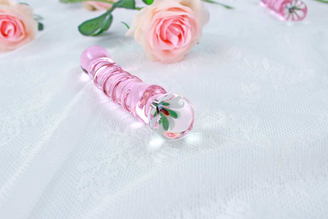 Pink glass ridged dildo with a flower base laid on white lace with pink roses in the background.