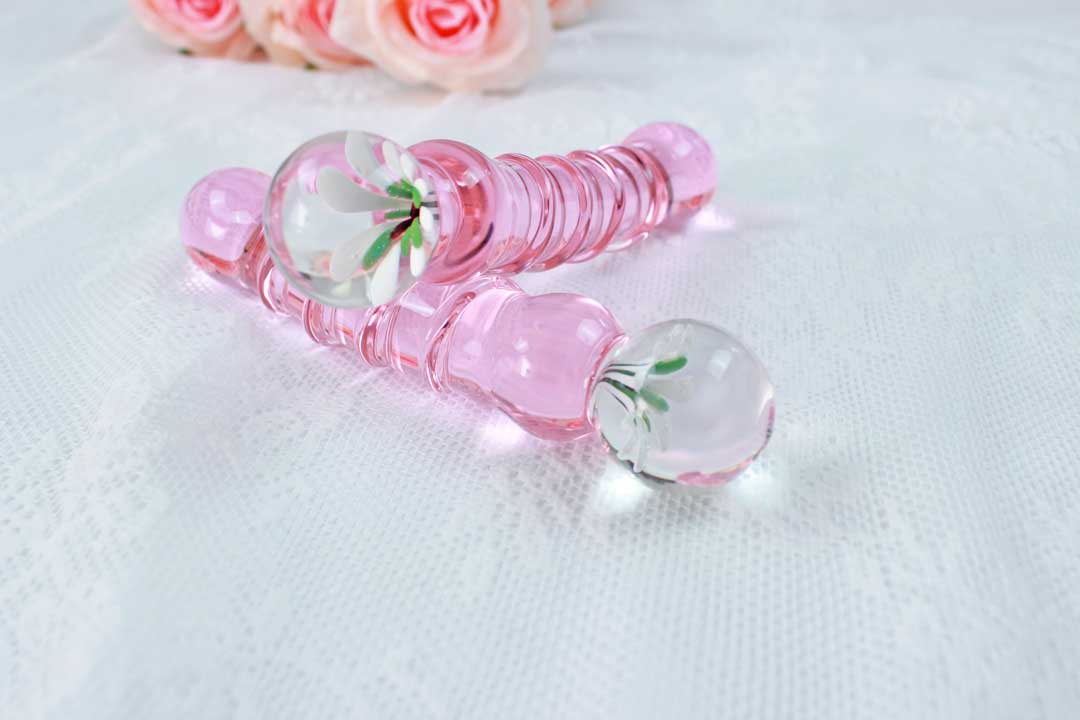 A glass pink ridged dildo with a flower base set on top of another glass pink ridged dildo with a flower base on a lace background.