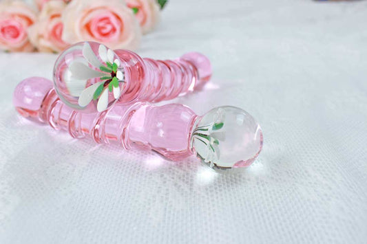 A glass pink ridged dildo with a flower base set on top of another glass pink ridged dildo with a flower base on a lace background.