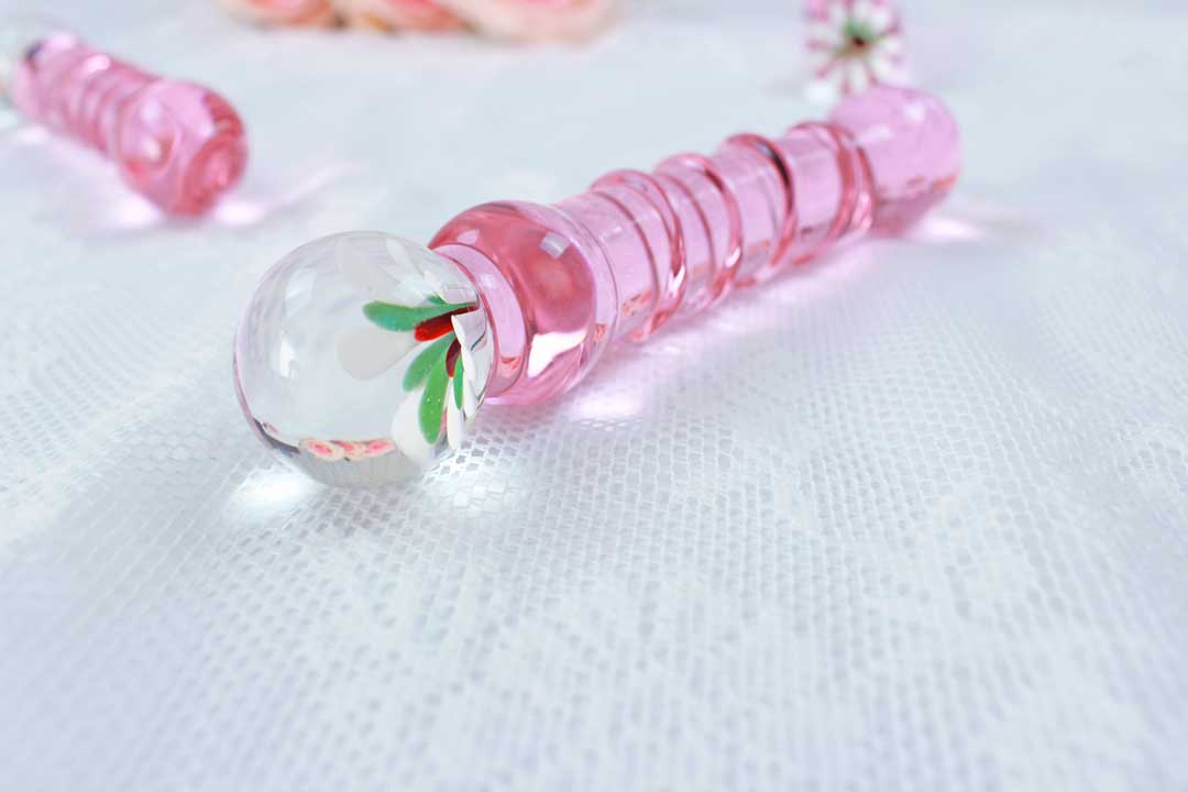 A pink glass dildo with a flower base laid out with another pink glass dildo in the background set on white lace.