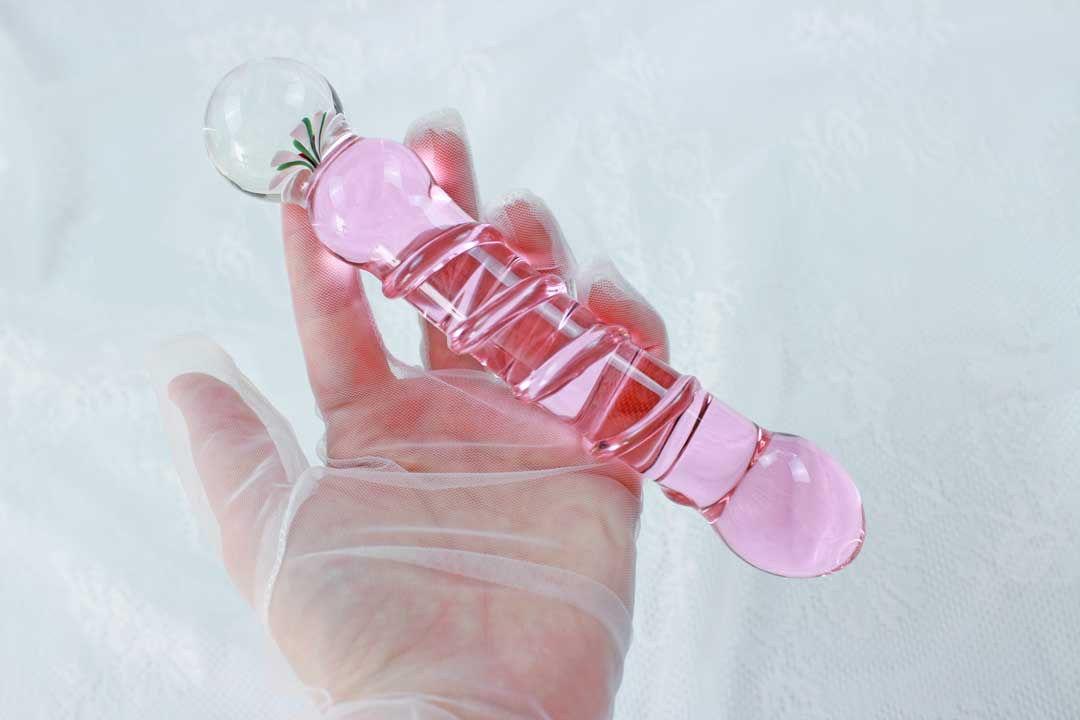 A pink glass ridged dildo with a flower base being held by a lace gloved hand over a lace backdrop.