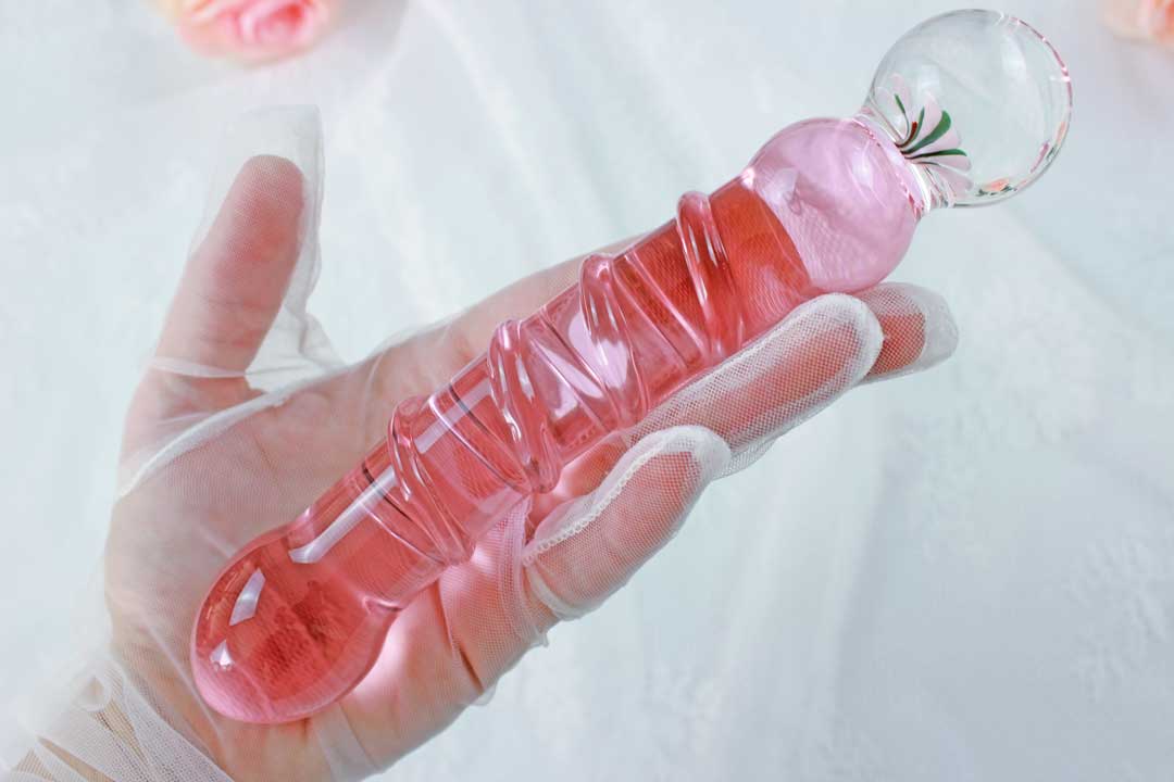 A pink glass ridged dildo with a flower base being held by a lace gloved hand over a lace backdrop.