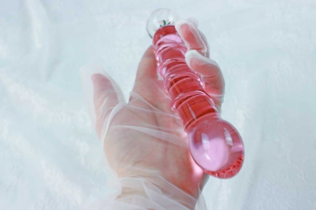 A pink glass ridged dildo with a flower base being held by a lace gloved hand over a lace backdrop.