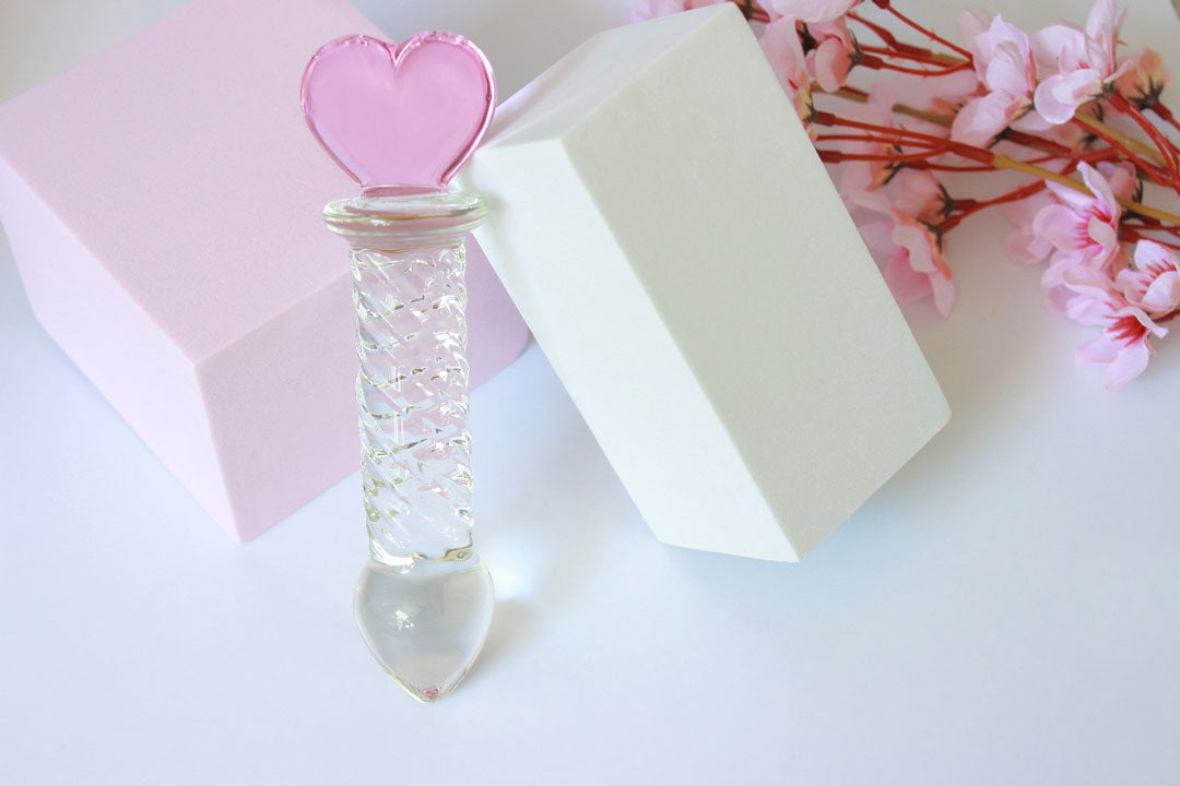 Glass ridged dildo with a clear shaft and a pink heart base leaning on a white and pink square elevated on a white background with flowers