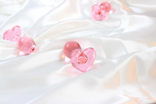 3 pink glass butt plugs with heart bases on white satin fabric