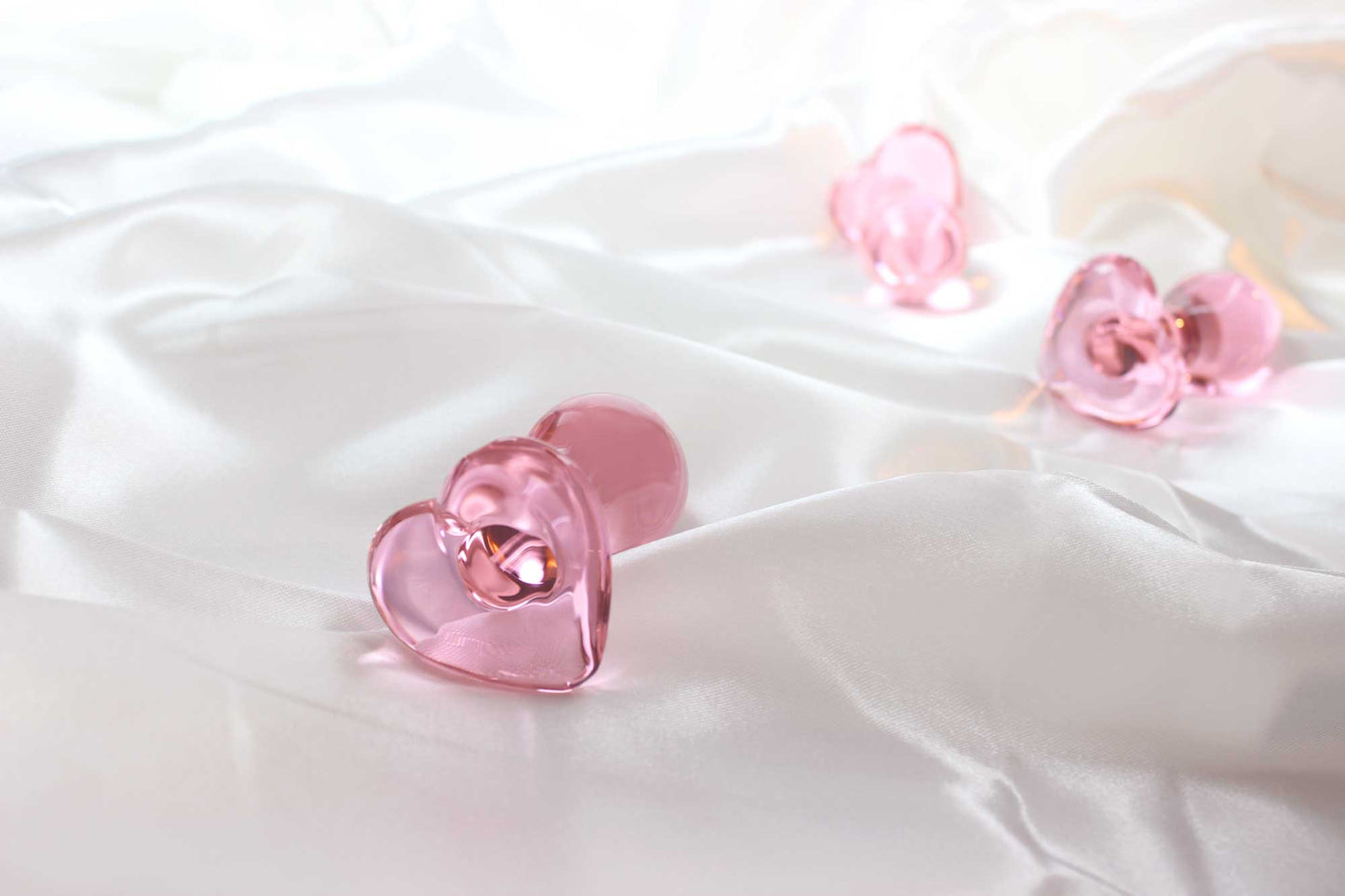 3 pink glass butt plugs with heart bases on white satin fabric