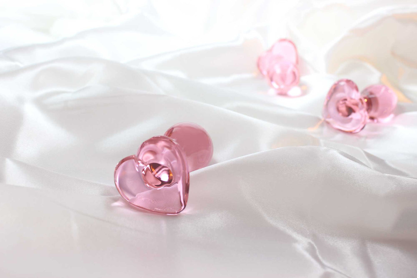 3 pink glass butt plugs with heart bases on white satin fabric