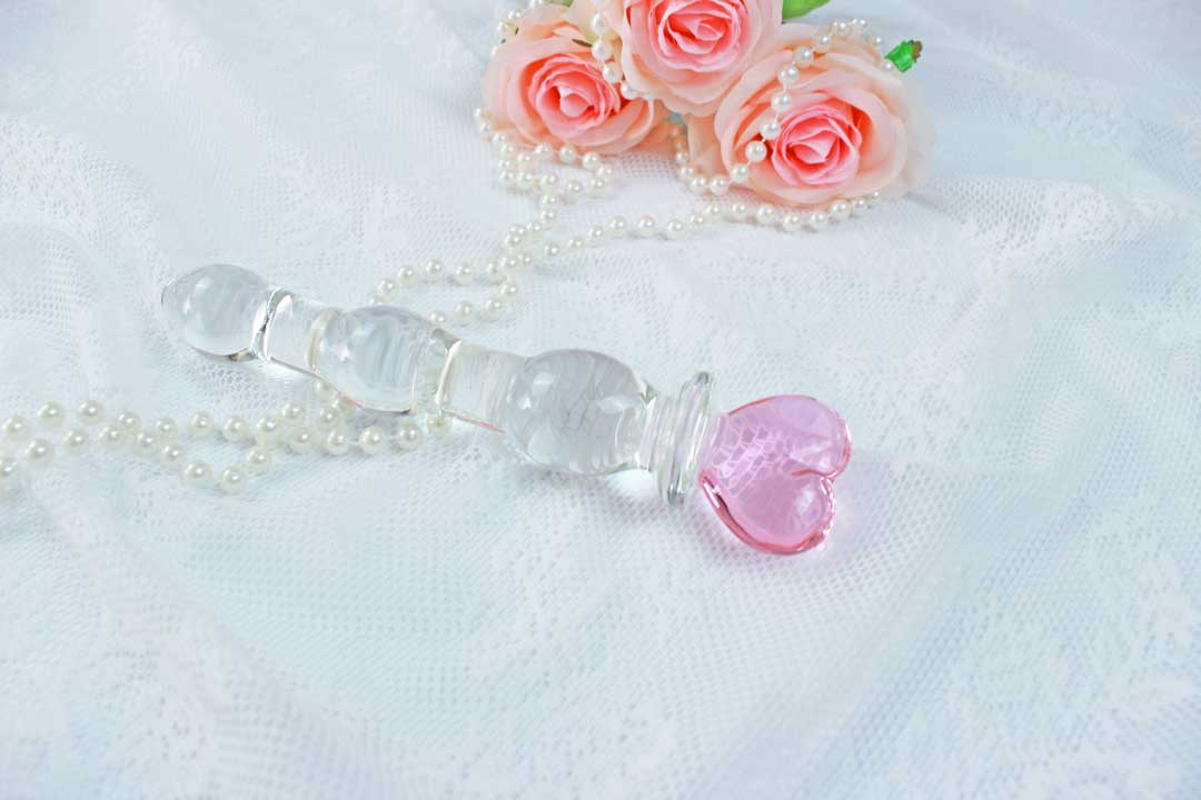 Knotted glass dildo with a pink heart base laid out on white pearl beads and lace with a bundle of pink roses in the background.