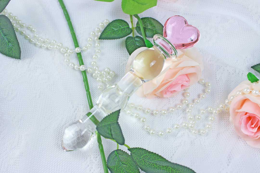 knotted glass dildo with a pink heart base laid on top of pink roses, pearl beads, and a white lace background.