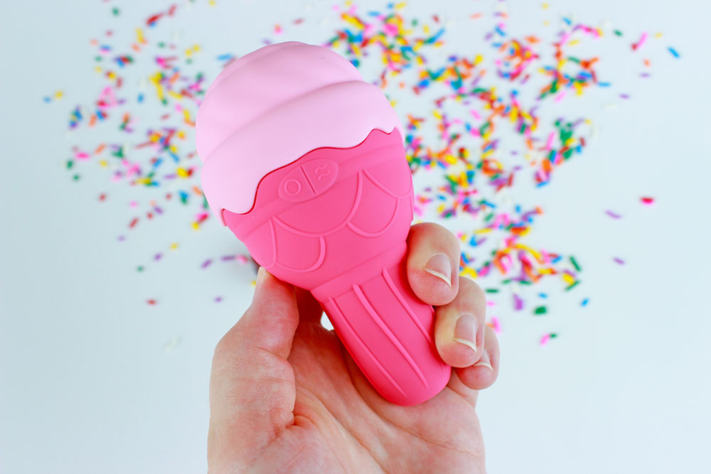 Hand holding a pink silicone ice cream shaped vibrator above a white background with scattered rainbow sprinkles
