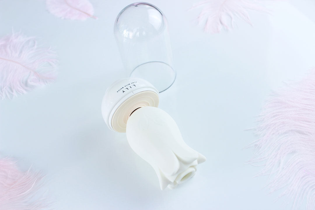 White lily suction vibrator laying in front of its clear plastic lid on a white background surrounded by pink feathers