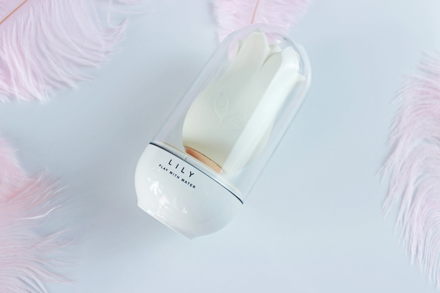 White lily suction vibrator in clear plastic case on a white background surrounded by pink feathers