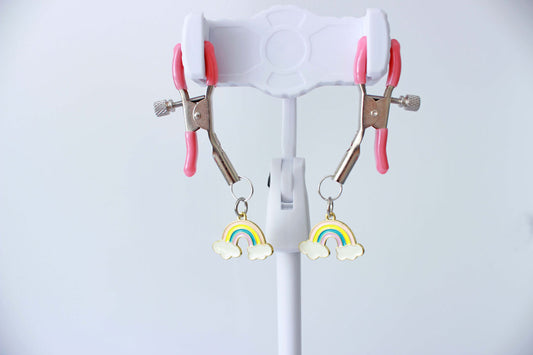 Silver adjustable tension screw nipple clamps with pink grip caps and charms of rainbows hanging from each clamp held up on a jewelry stand with a white background.