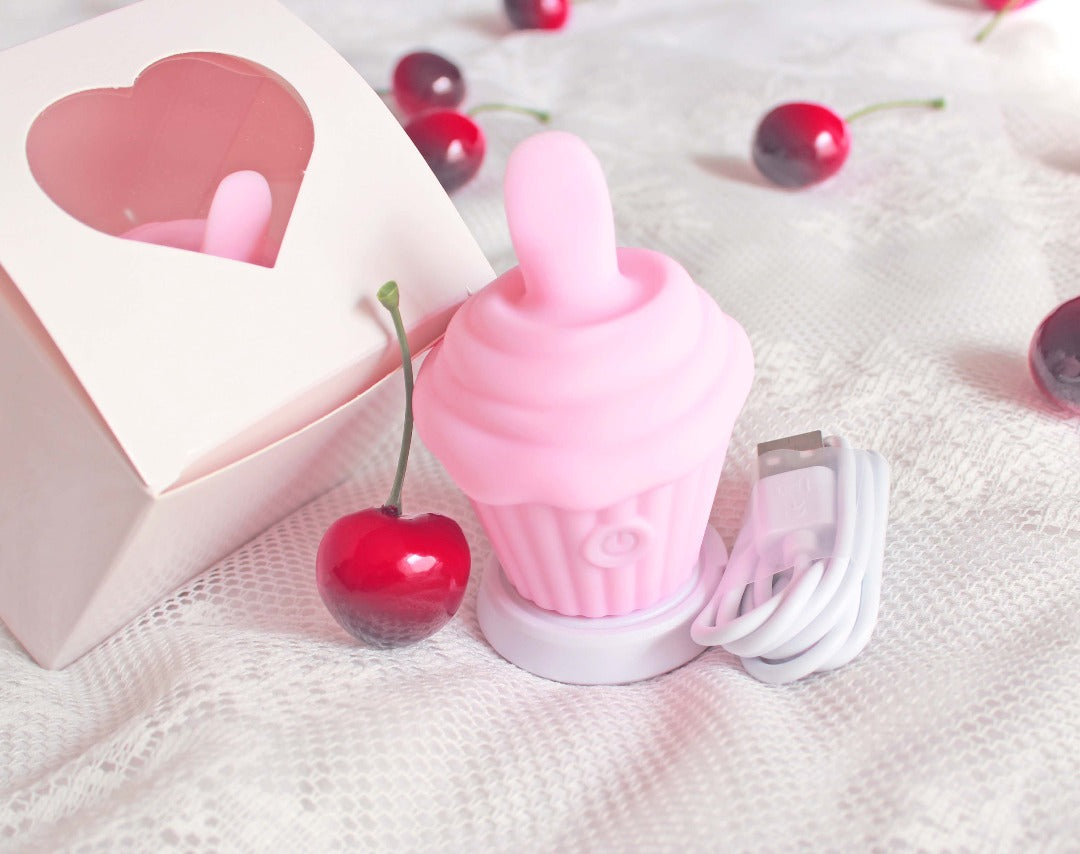 Pink cupcake licking vibrator upright on charging stand next to USB charging cord and box with heart cutout, surrounded by cherries