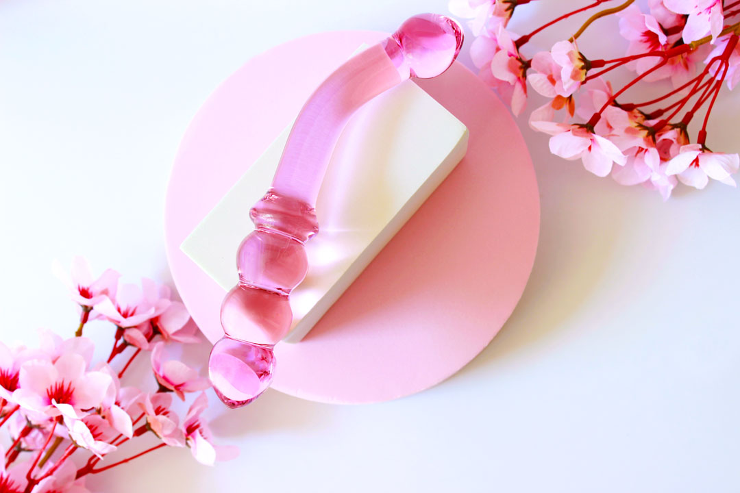 Pink glass double ended dildo with ribbing on one side on an elevated white square on a white background with flowers.