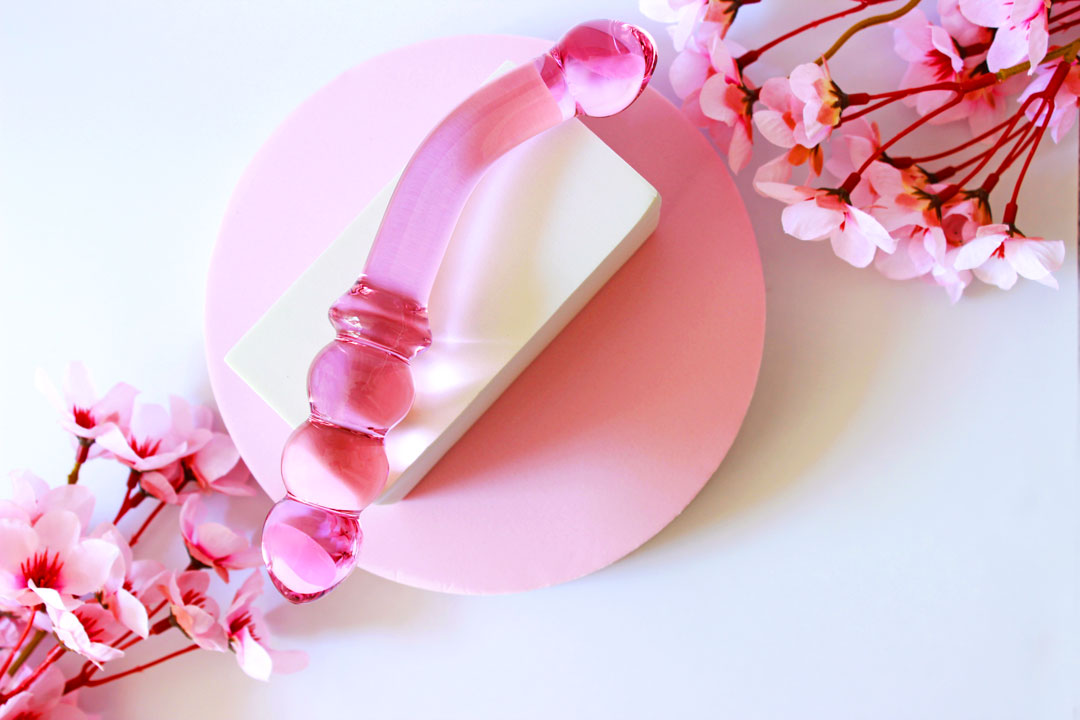 Pink glass double ended dildo with ribbing on one side on an elevated white square on a white background with flowers.