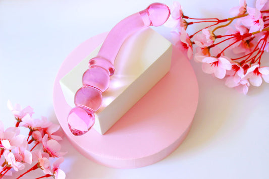 Pink glass double ended dildo with ribbing on one side on an elevated white square on a white background with flowers.