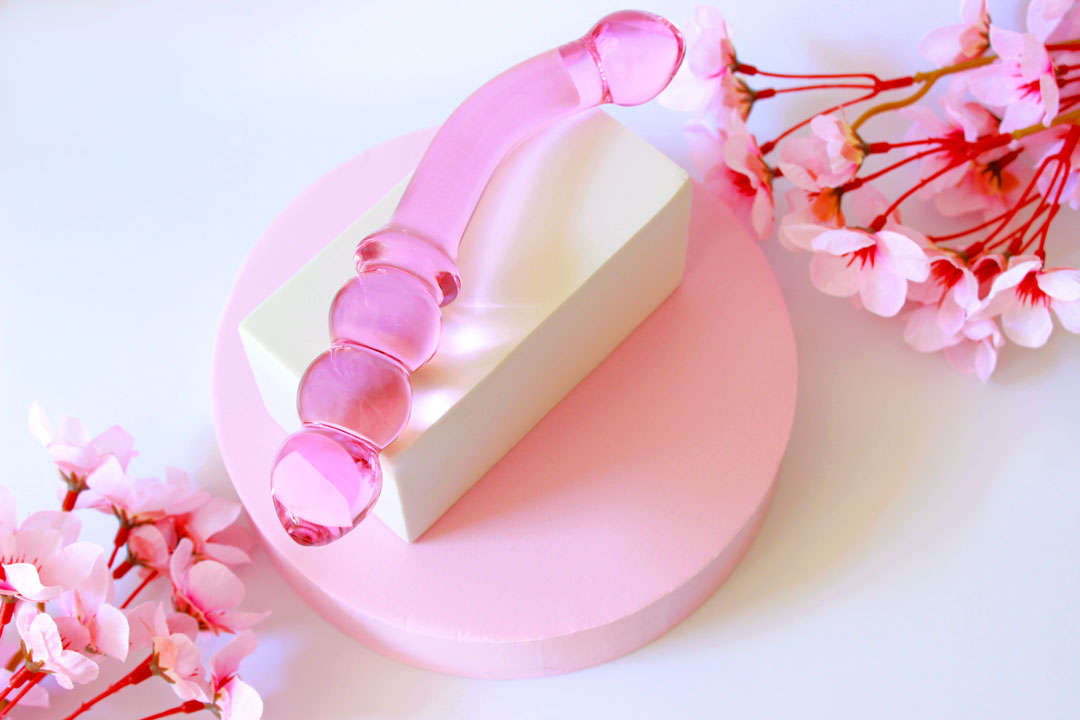 Pink glass double ended dildo with ribbing on one side on an elevated white square on a white background with flowers.