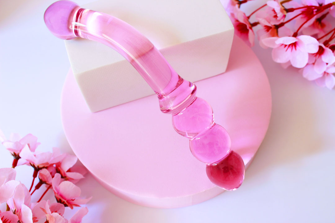 Pink glass double ended dildo with ribbing on one side on an elevated white square on a white background with flowers.