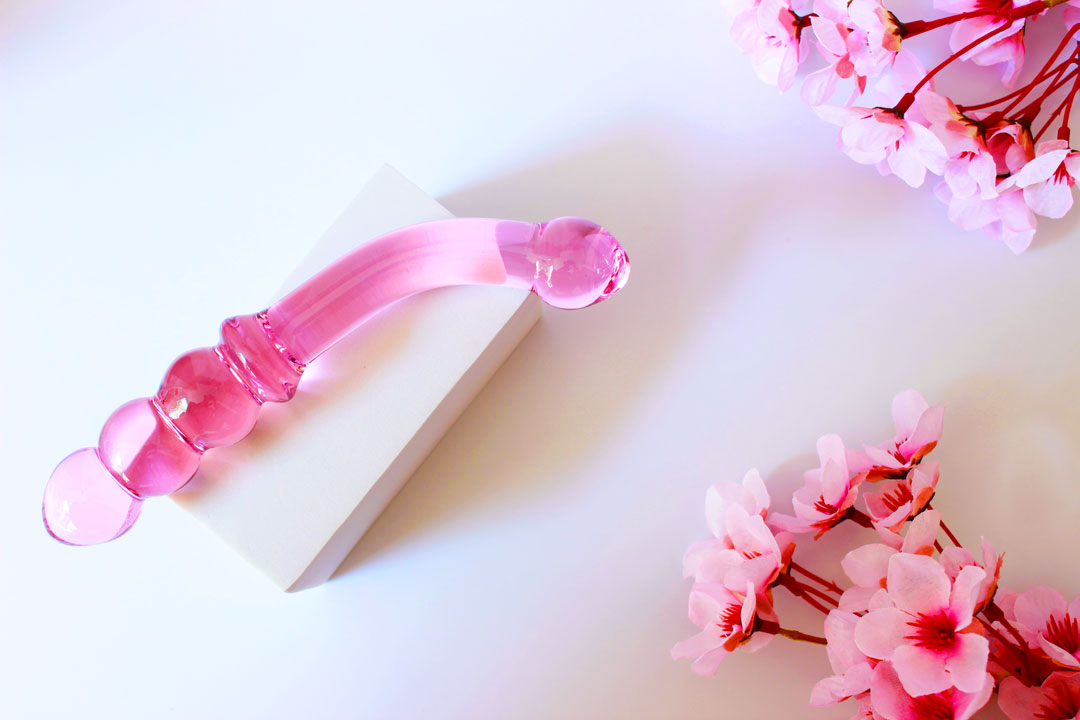 Pink glass double ended dildo with ribbing on one side on an elevated white square on a white background with flowers.