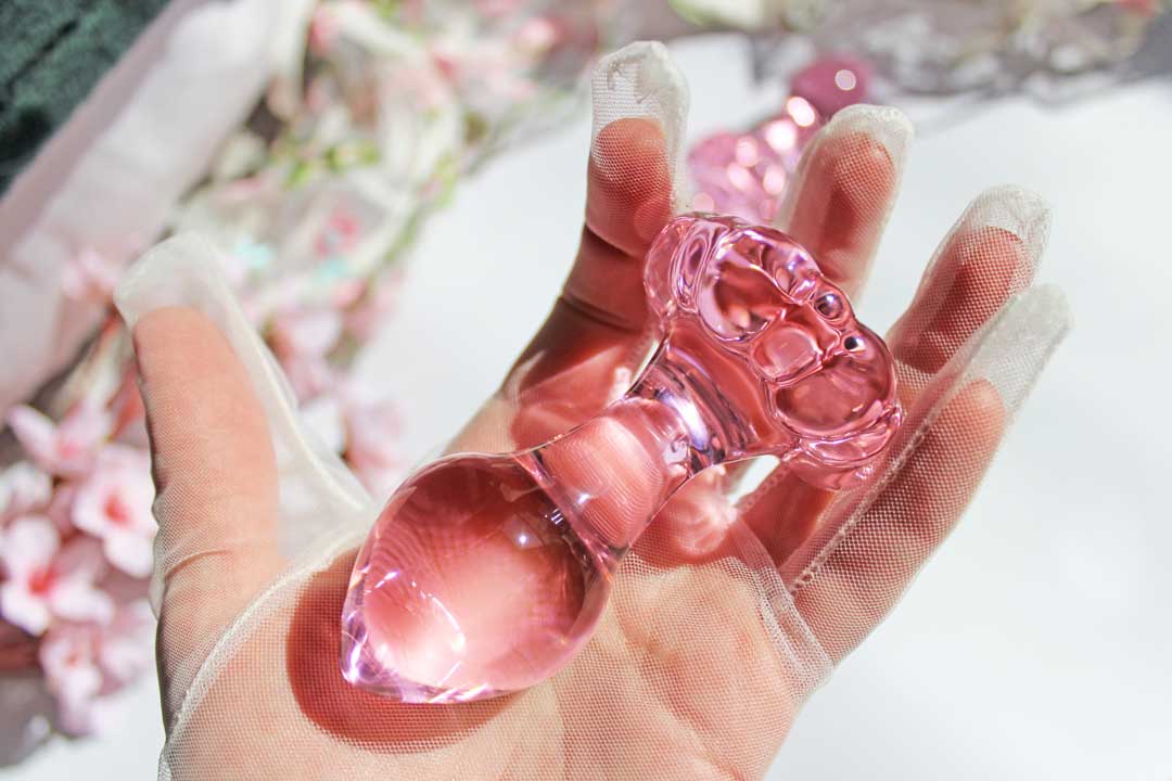 A pink glass daisy butt plug being held in a gloved hand over sheets and flowers.
