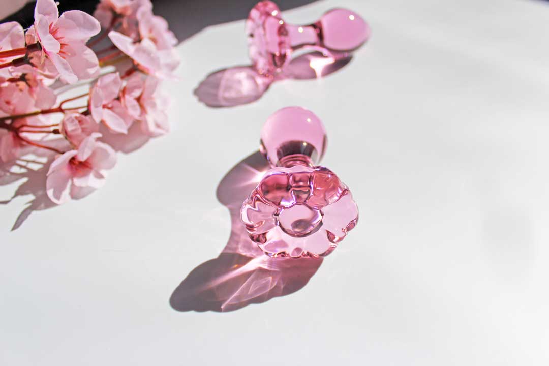 Two glass pink daisy butt plugs laid out on a white background with flowers.