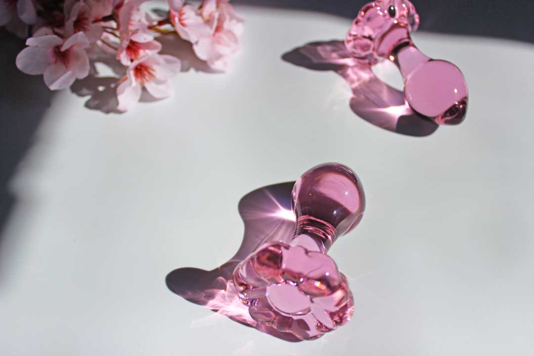 Two glass pink daisy butt plugs laid out on a white background with flowers.