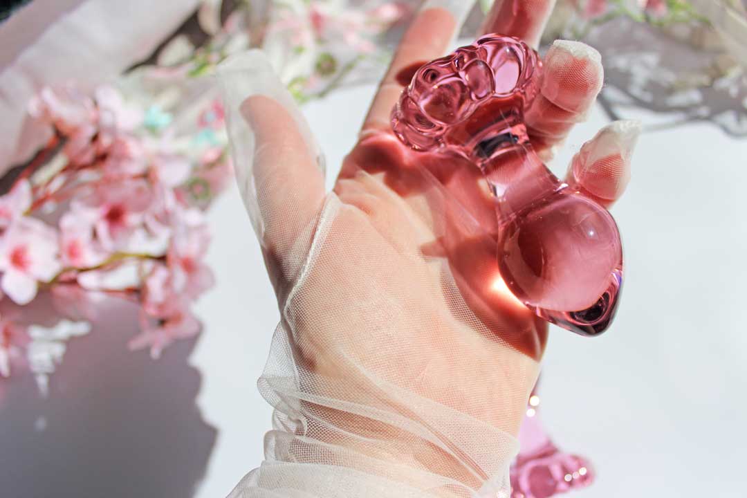 A pink glass daisy butt plug being held in a gloved hand over sheets and flowers.