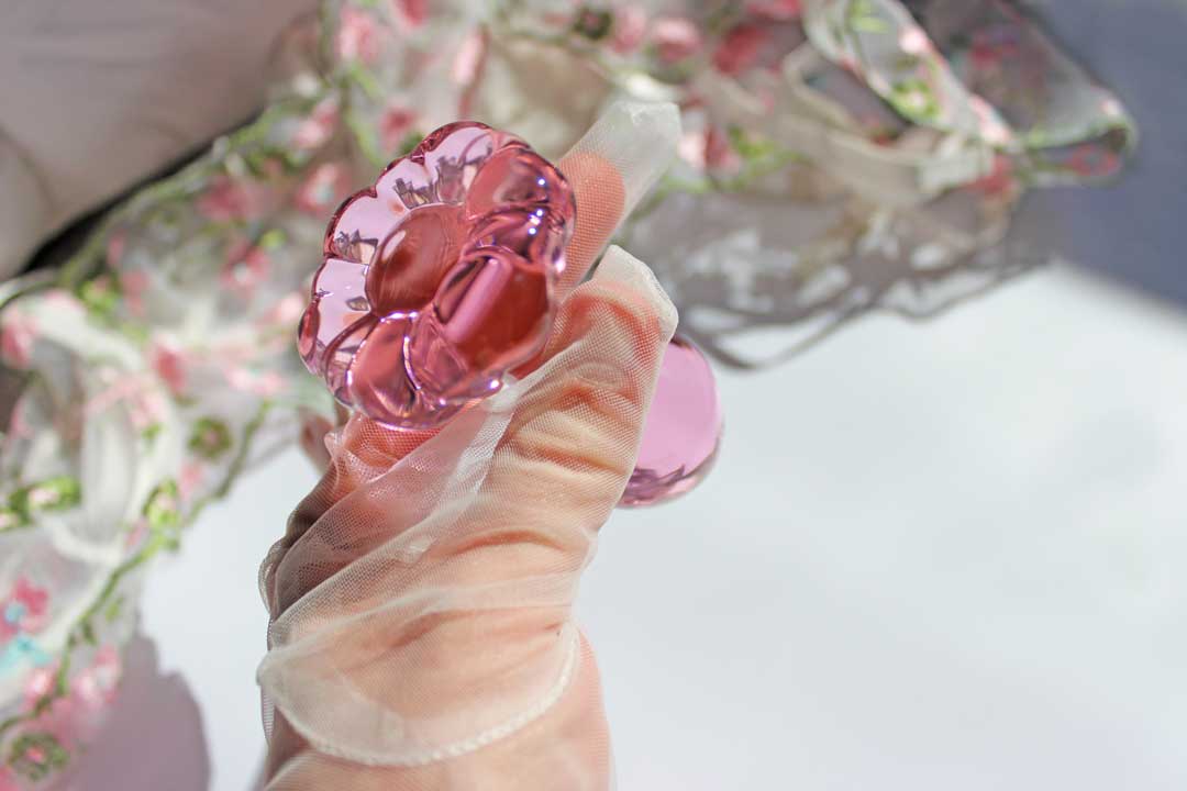 A pink glass daisy butt plug being held in a gloved hand over sheets and flowers.