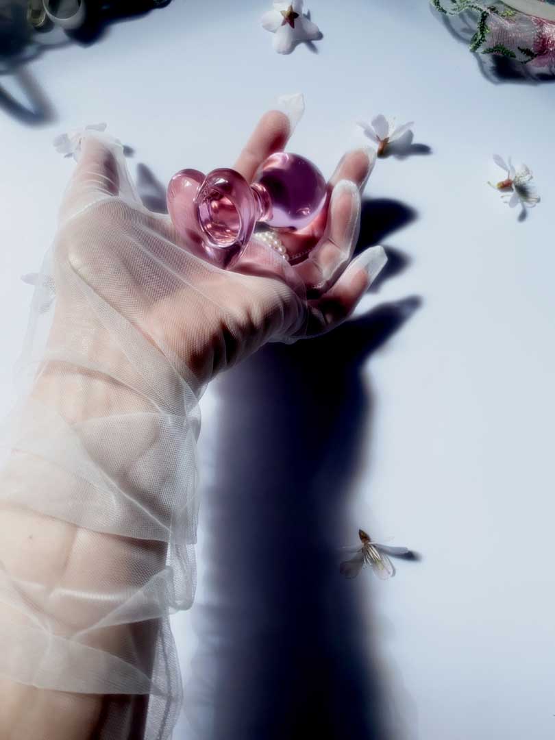 Glass pink butt plug with heart base being held in a gloved hand over a white background with flowers