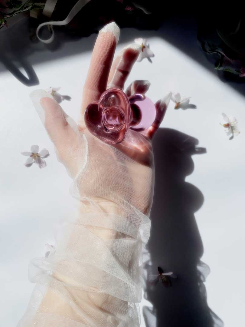 Glass pink butt plug with heart base being held in a gloved hand over a white background with flowers