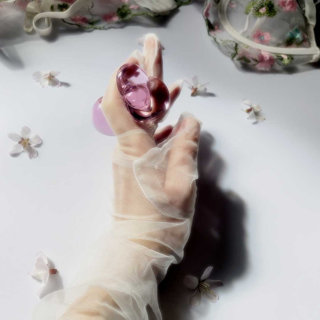 Glass pink butt plug with heart base being held in a gloved hand over a white background with flowers