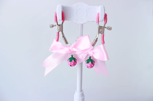 Silver adjustable screw nipple clamps with pink pvc caps and pink bows with pink strawberry bells hanging from them on a jewelry stand on a white background.