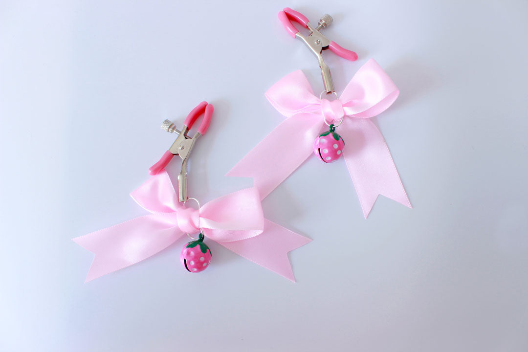 Silver ajustable screw nipple clamps with pink pvc cap ends and pink strawberry bells with pink bows hanging from them on a white background.