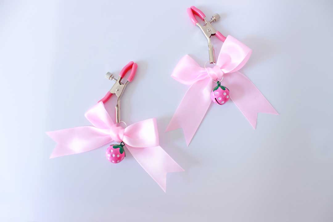 Silver ajustable screw nipple clamps with pink pvc cap ends and pink strawberry bells with pink bows hanging from them on a white background.