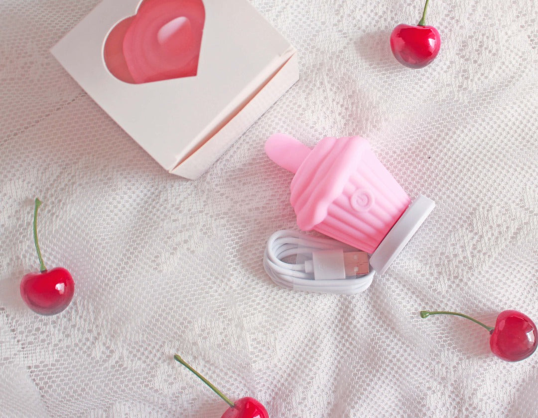 Pink cupcake licking vibrator laying next to USB charging cord and box with heart cutout, surrounded by cherries