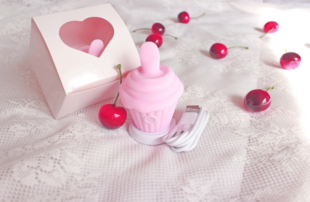 Pink cupcake licking vibrator upright on charging stand next to USB charging cord and box with heart cutout, surrounded by cherries