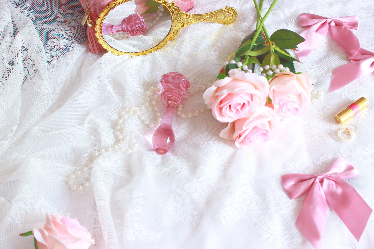 A glass rose plug and rose flowers being reflected into a golden hand mirror on a white lace sheet.