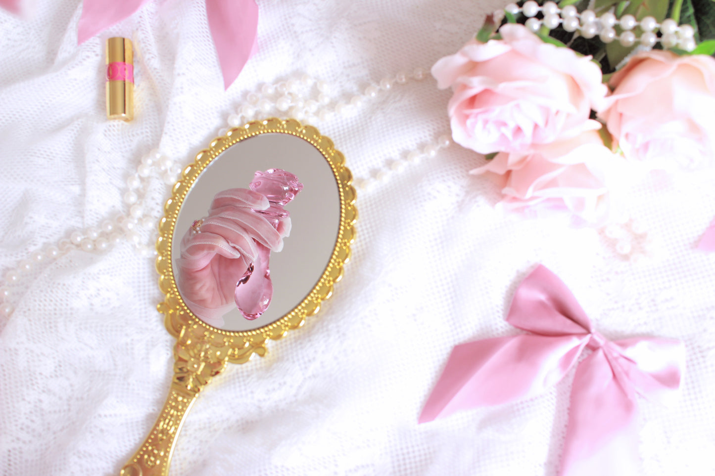 Pink glass rose plug being held in up in front of a gold mirror placed on a lace sheet with pearls, pink flowers, and bows.