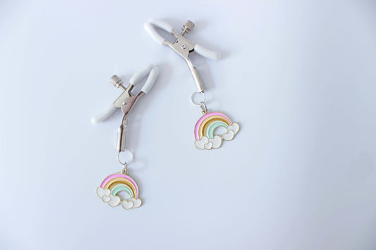 Silver adjustable tension screw nipple clamps with white grip caps and charms of rainbows hanging from each clamp set on a plain white background.