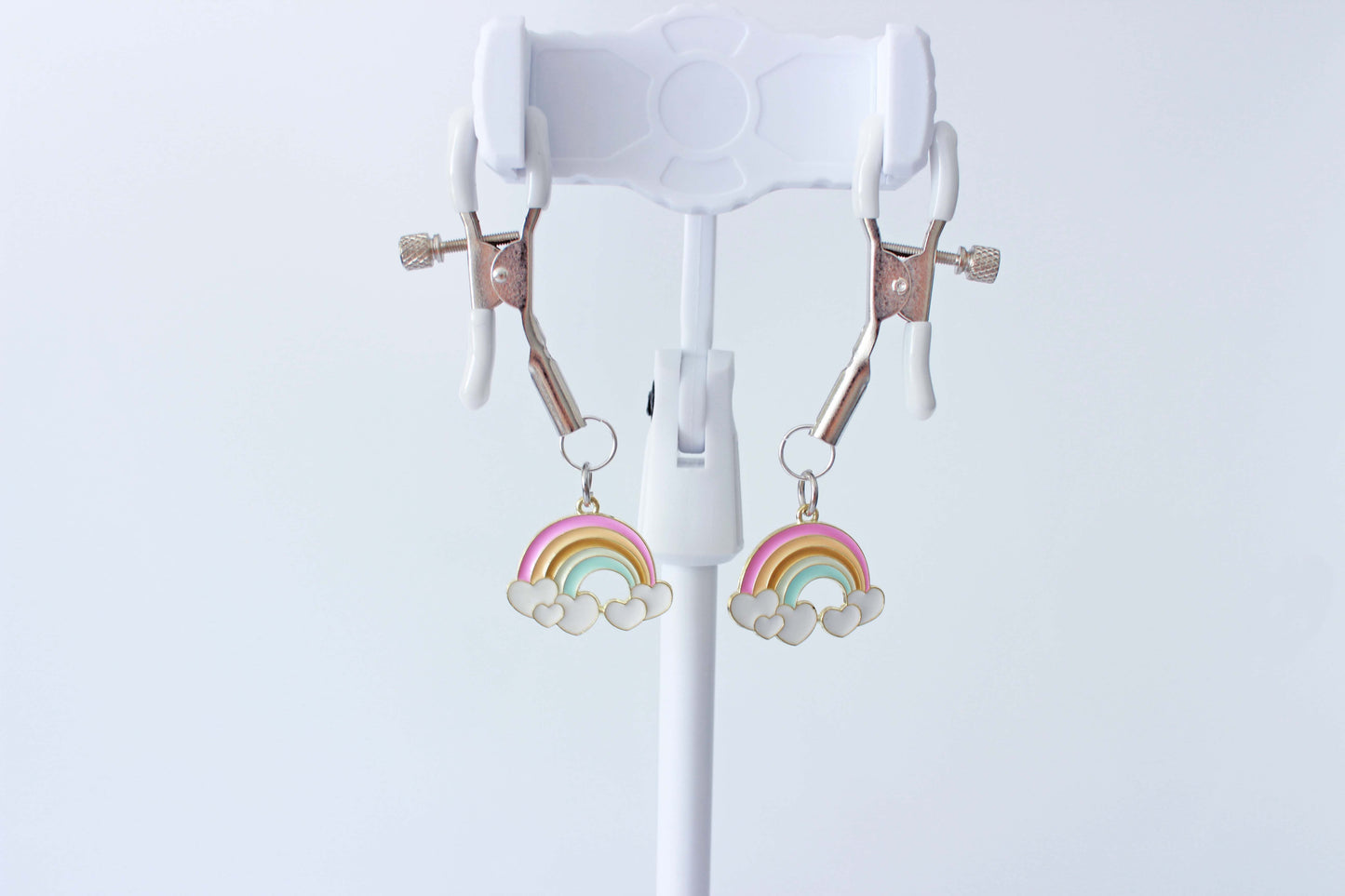 Silver adjustable tension screw nipple clamps with white grip caps and charms of rainbows hanging from each clamp held up on a jewlery stand with a white background.