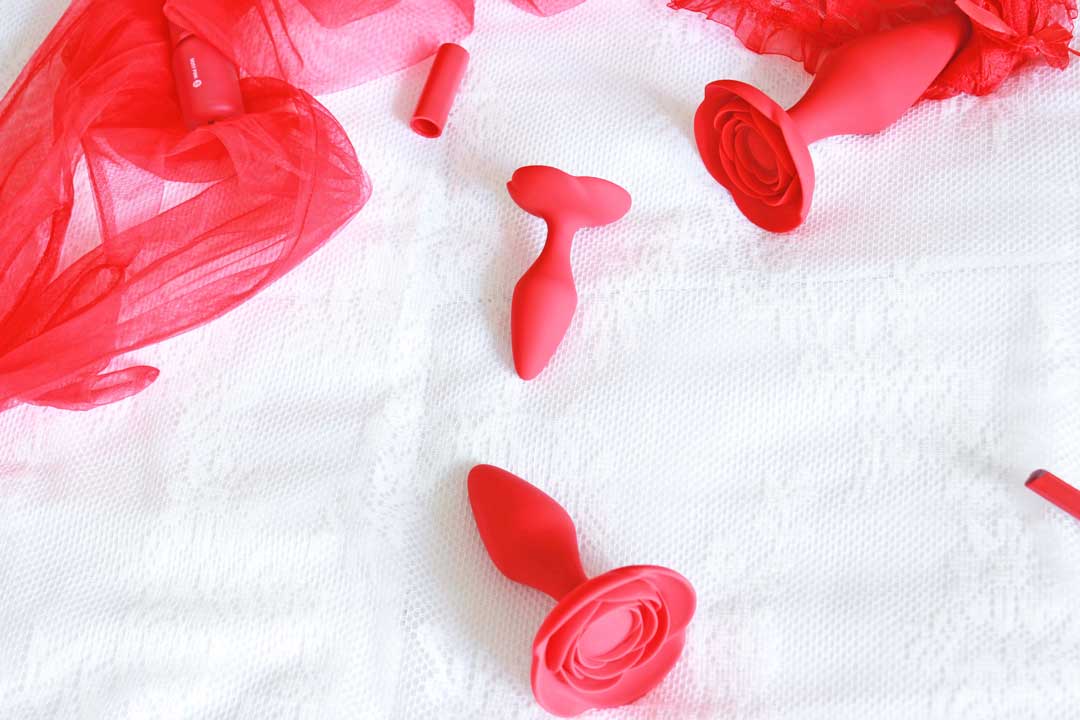 3 red rose anal training butt plugs on a white lace background surrounded by various red items